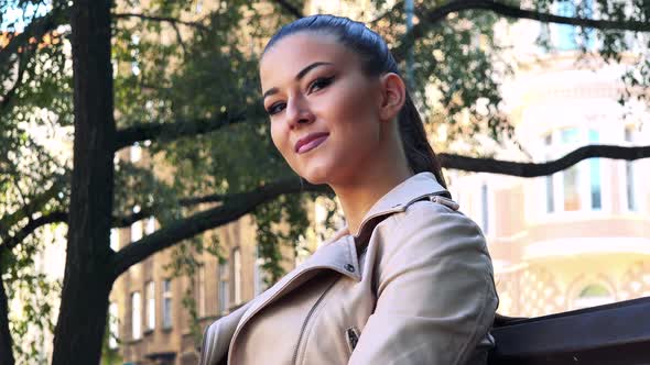 Young Beautiful Woman Sits on the Bench in Park and Looks Around - Closeup alt
