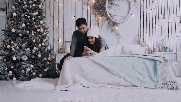 Young Couple Sitting on the Bed Next To the Christmas Tree Watching the News on the Phone alt