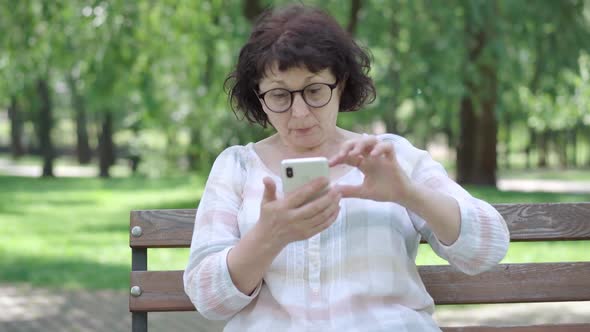 Senior Woman with Poor Eyesight Looking at Smartphone Screen in Sunny Park. Portrait of Female alt