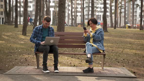 Woman and man sitting in park on bench, working on laptop and communicating alt