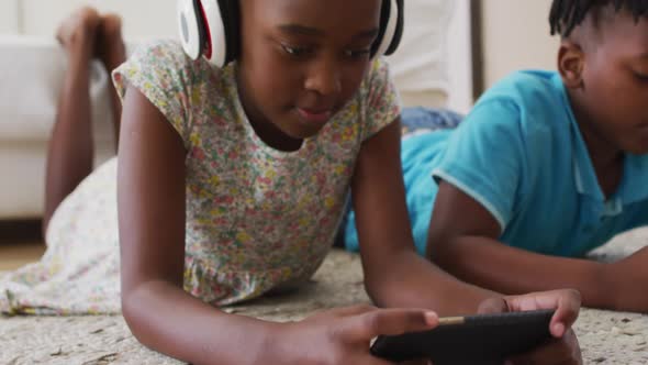 African american brother and sister using electronic devices lying on the floor at home alt