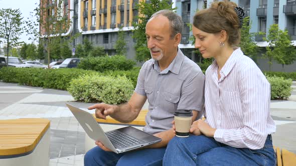 Young Employee is Teaching Her Older Colleague How to Use Laptop and Corporate Software Sitting on alt