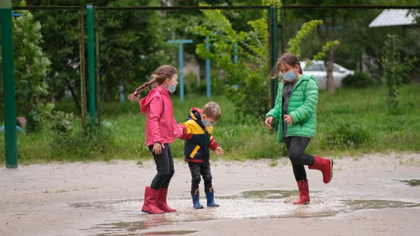 Two little girls and a boy in medical masks and rubber boots jump through puddles 