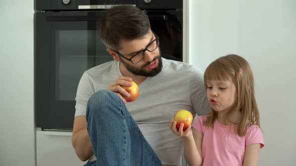 Happy Family Smiling Father and Little Daughter Sit on Floor at Home and Eat Ripe Apples alt