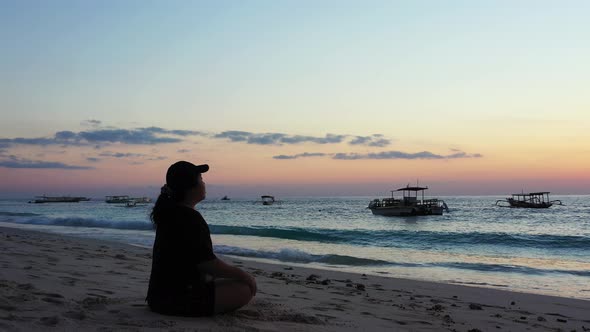 Sporty woman with a hat meditation in the early morning on the peaceful sandy beach. Boats waving on alt