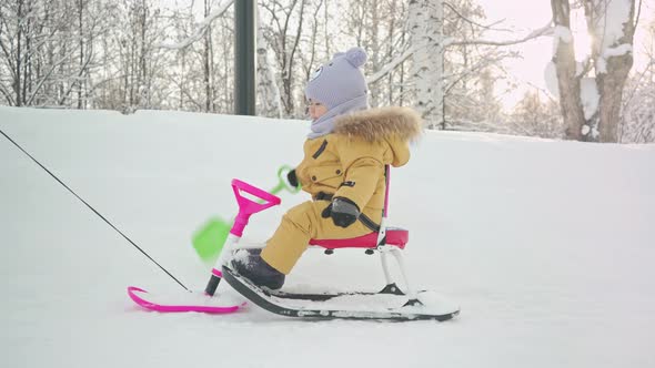A kid rides a snowcat sled with a shovel toy in his hand along a snowy path in a city park. alt