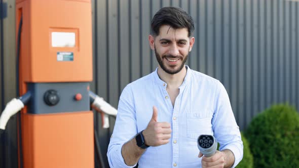 Bearded Hipster Man Standing Near Electric Charging Station Looking at Camera and Showing Thumb Up