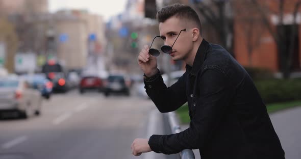Young Man Leaning on Barriers Looks at the City Car Traffic alt