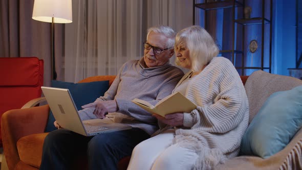 Senior Grandparents Couple Reading Book Using Laptop Pc on Couch in Night Living Room at Home alt