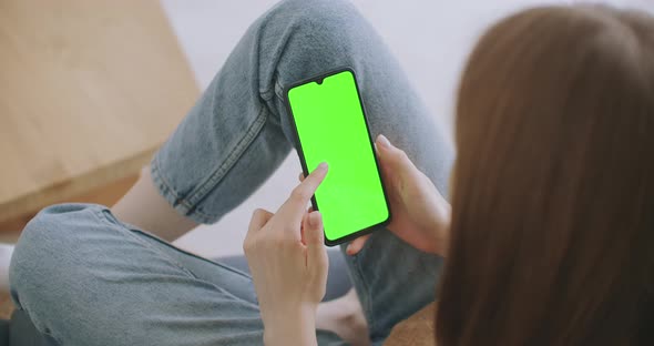 Point of View of Woman at Modern Room Sitting on a Chair Using Phone With Green Mock-up Screen alt