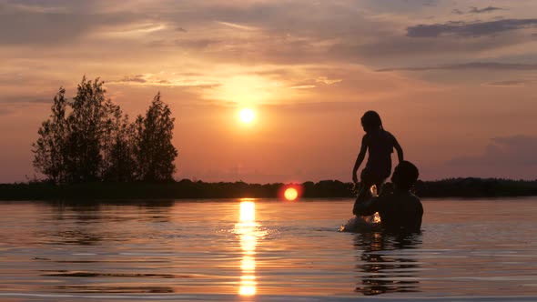 Father and son silhouette playing in water at sunset. Static shot. alt