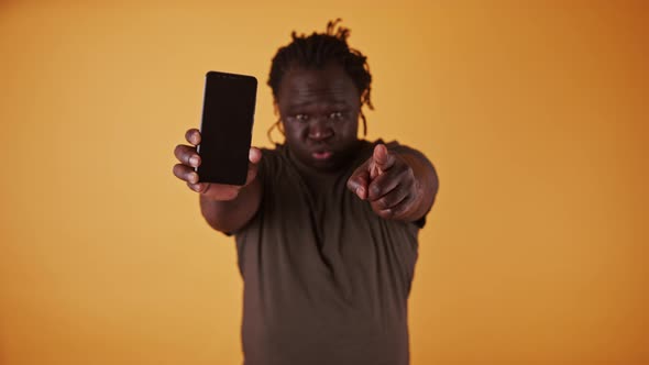 Angry African Man Holding Smartphone and Pointing Fingers Towards Camera alt