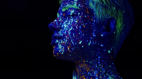 Man Face Painted with Fluorescent Colors in Profile Turns Head and Looks Into Camera alt