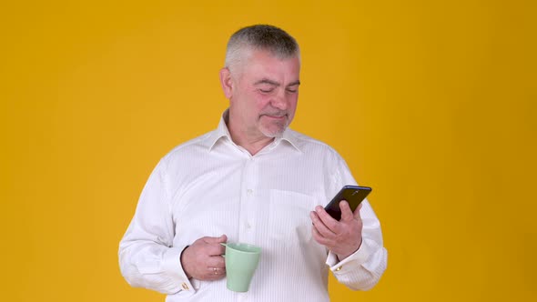 Handsome adult man with a mug of coffee or tea uses smartphone texting messages alt