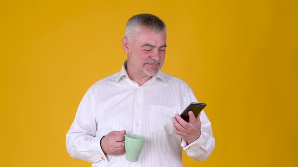 Handsome adult man with a mug of coffee or tea uses smartphone texting messages