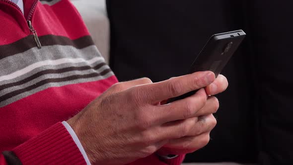 An Elderly Man Sits on a Couch in an Apartment and Works on a Smartphone - Closeup alt