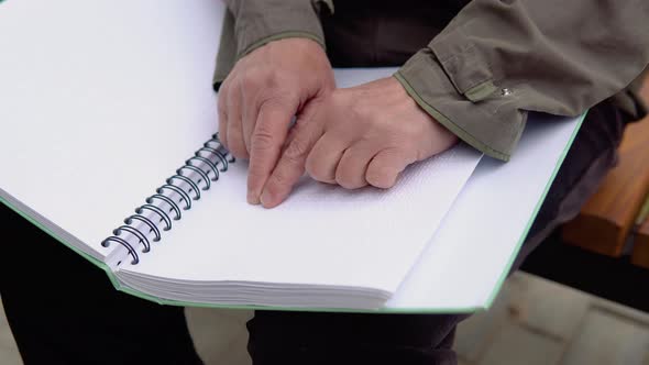 Senior Blind Man Sitting on Bench in City Park and Reading a Braille Book alt