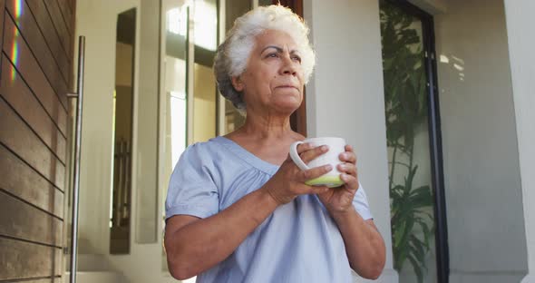 African american senior woman drinking coffee while standing at the front door at home alt