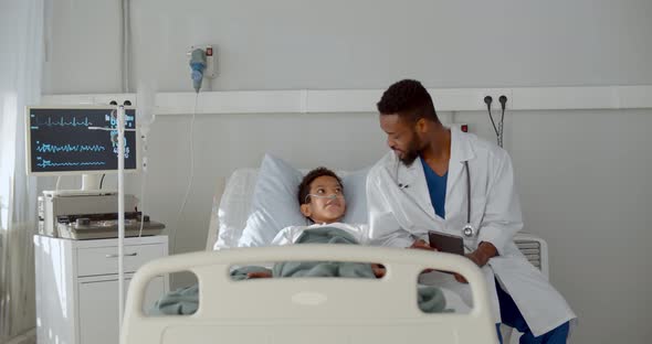 Young African Doctor Sitting on Hospital Bed and Talking To Preteen Boy Patient alt