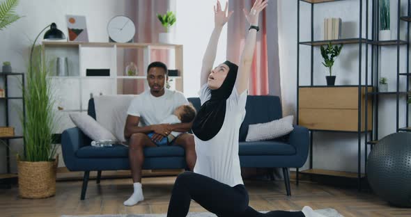 Woman in Headscarf Doing Stretching Exercises on the Floor near Her Black-Skinned Husband alt