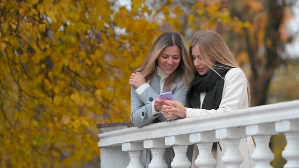 Two Happy Female Friends Using Mobile Phone In Autumn Park