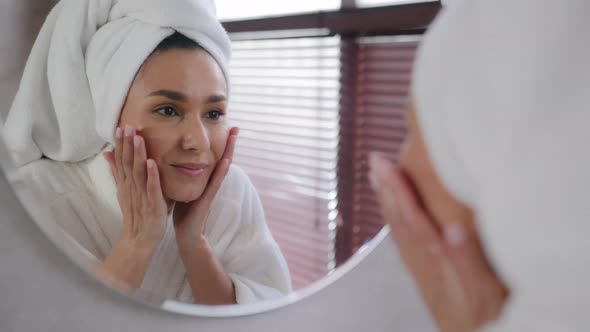 Young Hispanic Woman with Towel on Head After Shower Looks in Mirror Washes Face Healthy Soft Clean alt