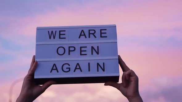 Hands Are Holding a Lightbox with a Sign We Are Open Again, Pink Blue Sky on the Background. We're