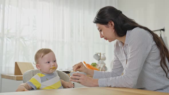 Caucasian attractive parents mother feeding healthy foods to baby boy toddler in kitchen at home. alt