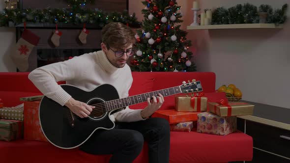Man is playing a merry song on guitar near decorated Christmas tree.