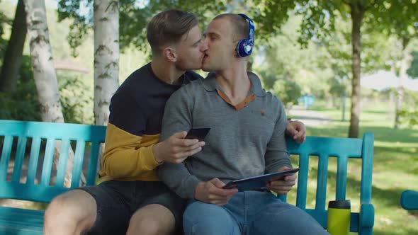 Cheerful Gay Couple with Gadgets Relaxing in Park alt