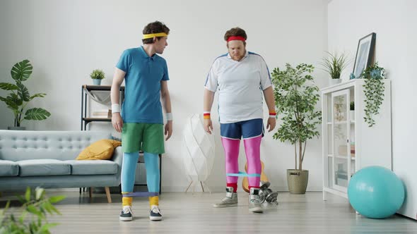 Slow Motion of Silly Guy Exercising with Stretch Band Kicking Friend Having Fun Indoors in Apartment alt