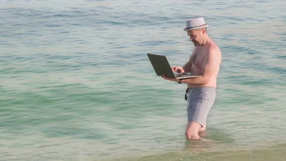 Male Freelancer in the Trunks and Hat Working Remotely at Vacation From the Sea