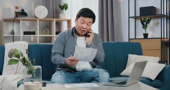 Chinese Male Worker Talking on Phone, Reading Report and Working on Laptop alt