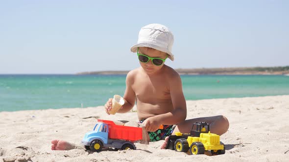 Child, Boy Playing with a Plastic Car in the Sand on the Beach and Eating Ice Cream. Hot Summer Day alt