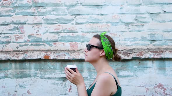 Young woman with bandana on hair standing on turquoise brick wall background with coffee to go city alt