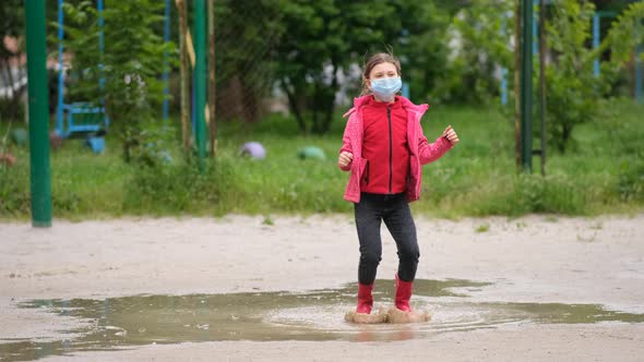 A little girl in a medical mask and rubber boots jumps through puddles on a spring day after rain.