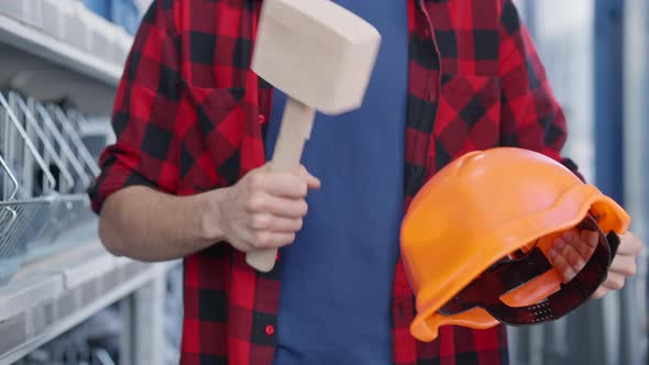 Unrecognizable Young Man Hitting Hard Hat with Hammer Standing at Shelves in Hardware Store alt