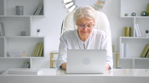 Aged businesswoman with glasses types on grey laptop with cheerful smile alt