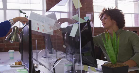 Diverse male and female business colleagues sitting at desks passing document over sneeze guard alt