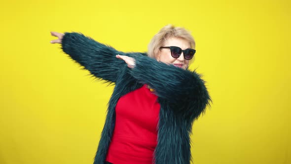 Stylish Senior Caucasian Woman in Fur Coat and Sunglasses Stretching Hands Aside at Yellow alt