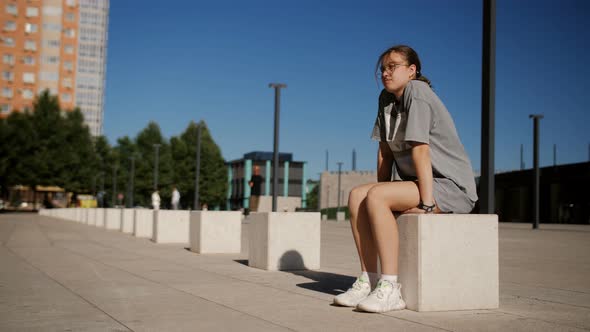 A Teenage Girl is Sitting on a Marble Cube in Front of a City Park on an Alley Waiting for Friends