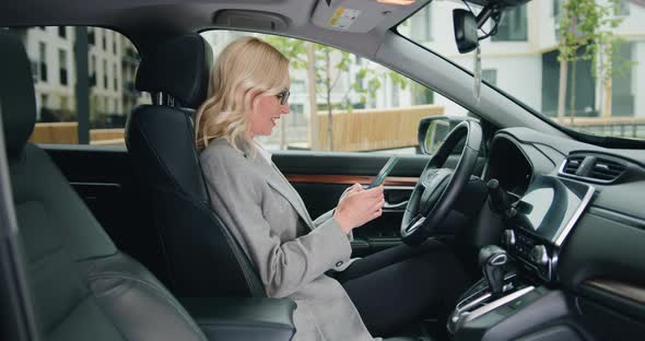 Smiling Adult Woman Typing Message on Smartphone while Sitting in Car on Drivers Seat. alt