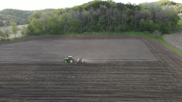 Farmer fertilizing and seeding smaller field at bottom of hill covered in trees in midwest.