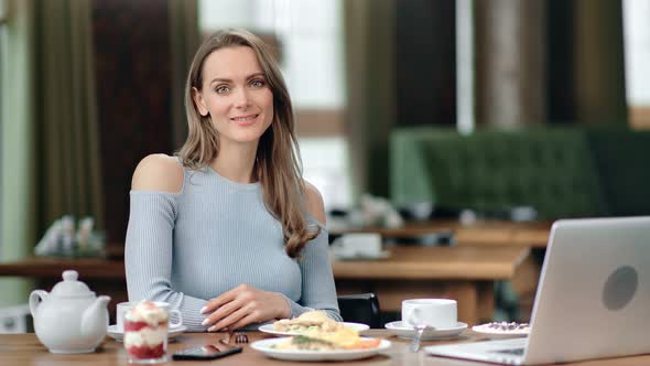 Portrait of Friendly Smiling Young Blonde Woman Posing Sitting at Table alt