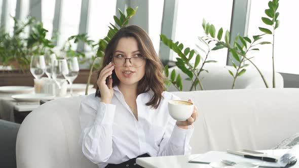 Pretty Businesswoman Holding Cup of Coffee and Looking at Screen of Mobile Phone Chatting