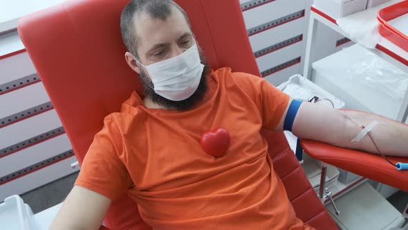 A Donor with a Beard Lies on a Couch with a Red Rubber Heart on His Chest. Man Donates Venous Blood alt