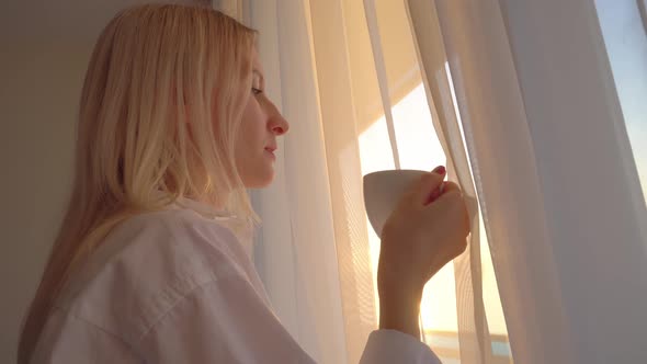 blonde in a white shirt looks out window, drinks a hot drink of tea or coffee, alt