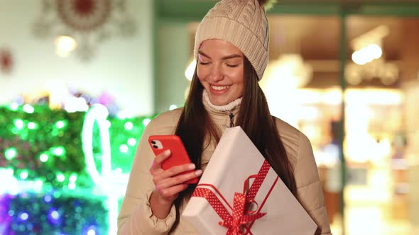 Portrait of Young Woman Stand Outdoors With Christmas Present Texting on Smartphone Smiling Happy