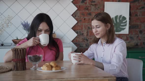 Two American Woman Drinking Coffee and Talking at Table in Kitchen Interior.