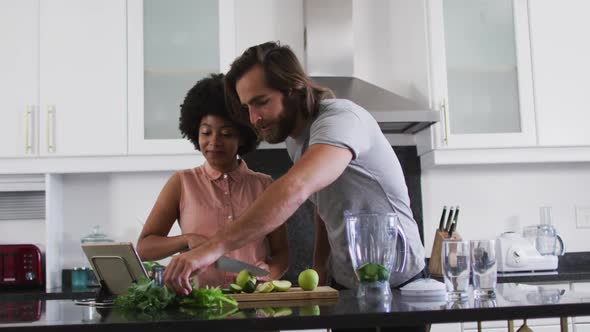Mixed race couple preparing vegetable smoothie while looking at digital tablet in the kitchen at hom alt